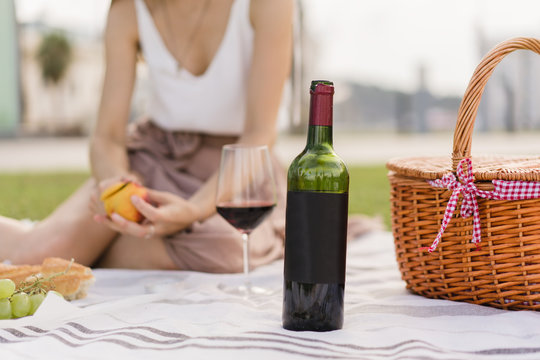Cropped image of a blank black bottle of red wine. Pretty woman having picnic in the park sitting on green grass and celebrating holidays with friends on the background. Space for you logo or design