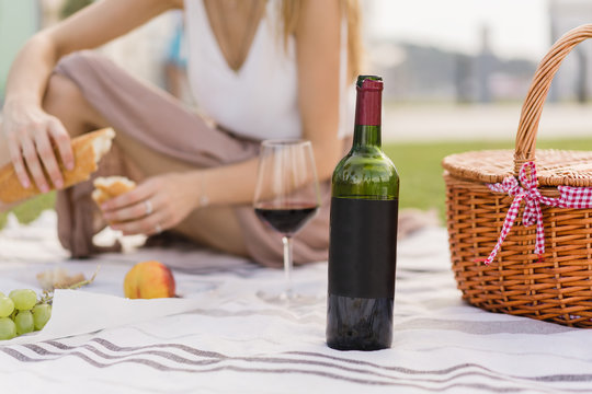 Close up of a blank bottle of red wine. Girl having picnic on the background. Space for you logo or design. Woman having lunch outdoors and enjoying holidays and weekends in the park with green grass