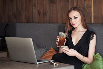 Young adorable woman with gorgeous eyes trendy makeup and stylish outfit drinking big glass cup of coffee with straw using notebook in cafe