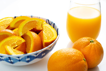 Freshly squeezed orange juice in a glass next to a bowl of orange slices and two oranges just picked off the tree on a white table for breakfast.