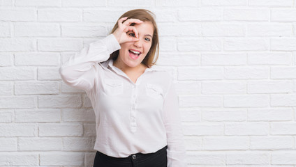 Young adult business woman over white brick wall with happy face smiling doing ok sign with hand on eye looking through fingers