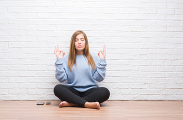 Young adult woman sitting on the floor in autumn over white brick wall relax and smiling with eyes closed doing meditation gesture with fingers. Yoga concept.