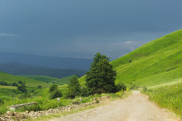 Obraz premium A rural road between the hills with trees along the edges and thunderstorm clouds in the summer