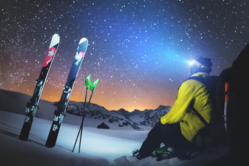 A skier sits at a stone in the mountains at night against a starry sky next to skis and sticks. The...