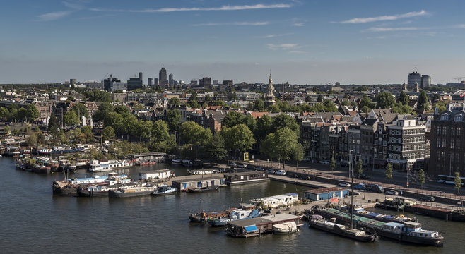 View north, over Amsterdam city, with many boats alongside the large canal. It is summertime and the sky is blue