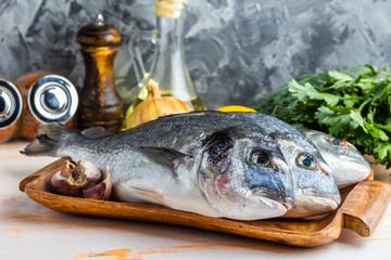 Three are ready to cook raw dorado fish on a wooden tray with herbs, lemon, onions, garlic, spices and olive oil on a gray textured background