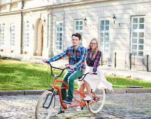Active romantic tourist couple, bearded man and smiling long-haired blond woman in white skirt riding tandem bike along paved city sidewalk on bright sunny day on blurred ancient building background.