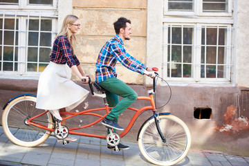 Young romantic pair, handsome man and pretty blond woman cycling together tandem double red bike along paved sidewalk on bright sunny summer day by old buildings - obrazy, fototapety, plakaty