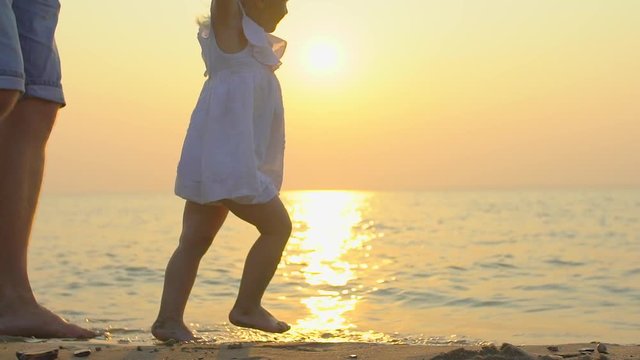 Little baby learns to walk. First Steps. Slow Motion 120 fps. Father is teaching him child, litle girl to do the first steps on a sand on the beach in summer. Baby feet. Close up of feet. Happy