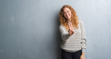 Fototapeta premium Young redhead woman over grey grunge wall smiling looking to the camera showing fingers doing victory sign. Number two.