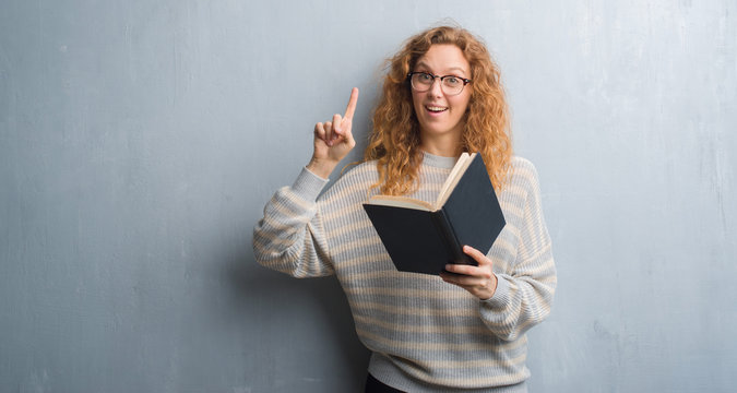 Young redhead woman over grey grunge wall reading a book surprised with an idea or question pointing finger with happy face, number one