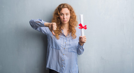 Young redhead business woman over grey grunge wall holding diploma with angry face, negative sign showing dislike with thumbs down, rejection concept