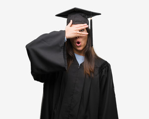Young hispanic woman wearing graduated cap and uniform peeking in shock covering face and eyes with hand, looking through fingers with embarrassed expression.