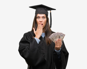 Young hispanic woman wearing graduated uniform holding dollars cover mouth with hand shocked with shame for mistake, expression of fear, scared in silence, secret concept
