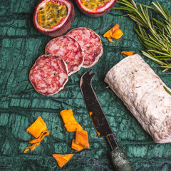 Christmas snack with dried sausage, passy fruit, dryed mango and green branches of fresh rosemary, overhead view on a cutting board of green stone.