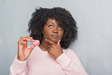 Young african american woman over grey grunge wall eating pink macaron serious face thinking about question, very confused idea