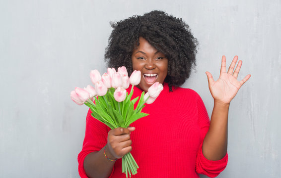 Young african american woman over grey grunge wall holding pink flowers bouquet very happy and excited, winner expression celebrating victory screaming with big smile and raised hands
