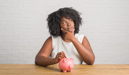Young african american woman sitting on the table holding piggy bank serious face thinking about...