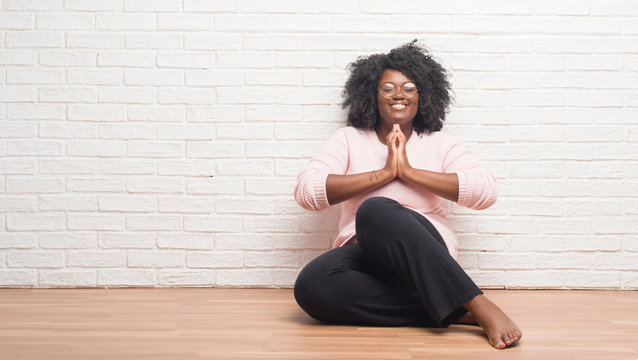 Young African American Woman Sitting On The Floor At Home Praying With Hands Together Asking For Forgiveness Smiling Confident.