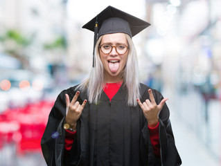 Young blonde woman wearing graduate uniform over isolated background shouting with crazy expression doing rock symbol with hands up. Music star. Heavy concept.