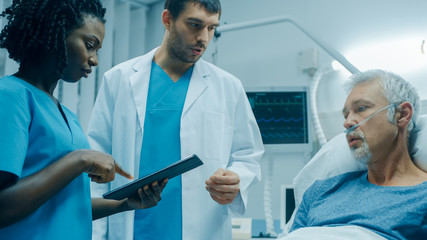 Obraz premium In the Hospital, Senior Man Lying in the Bed, Talks with Doctor and Nurse who Use Tablet Computer. Technology Helps Cure Patients, Modern Hospital Ward.