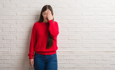 Young Chinese woman over brick wall covering one eye with hand with confident smile on face and surprise emotion.