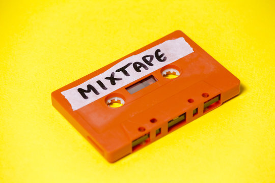 A vintage cassette tape (obsolete music technology), orange on a yellow surface, angled shot, carrying a label with the handwritten text Mixtape.
