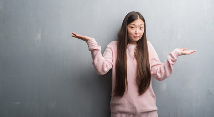 Young Chinese woman over grey wall clueless and confused expression with arms and hands raised....