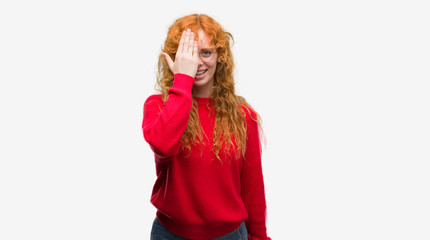 Young redhead woman wearing red sweater covering one eye with hand with confident smile on face and surprise emotion.