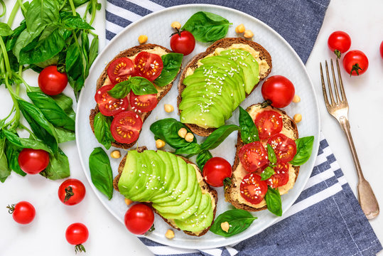 healthy avocado and tomatoes sandwiches with hummus, sesame and basil in a plate with towel and fork