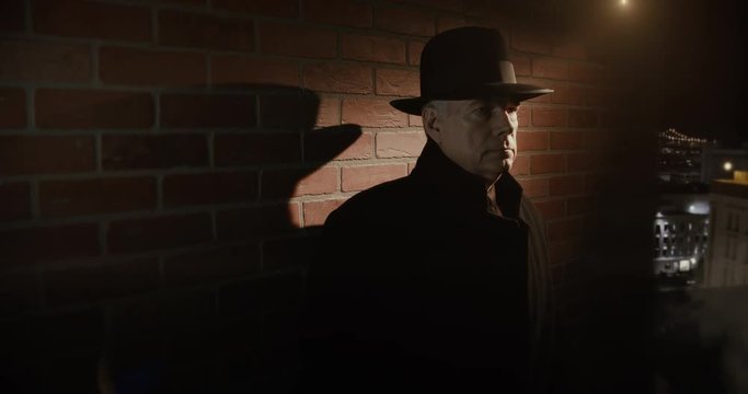 A classic film noir 1940s style scene with a suspicious looking character wearing a fedora who lights up a cigarette as he waits in the shadows of a dimly lit rooftop.