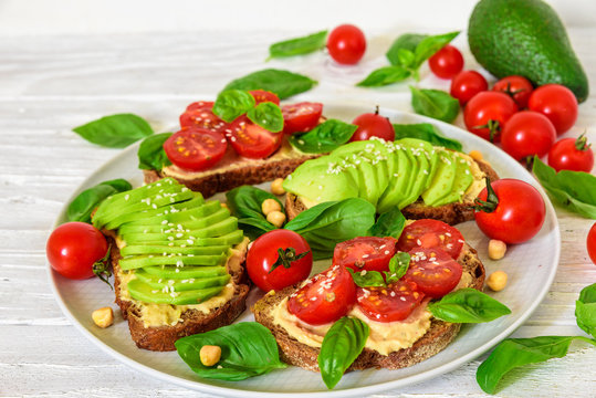 avocado and tomatoes sandwiches with hummus, sesame and basil in a plate over white wooden background