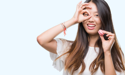 Young asian woman eating pink macaron sweet over isolated background with happy face smiling doing ok sign with hand on eye looking through fingers