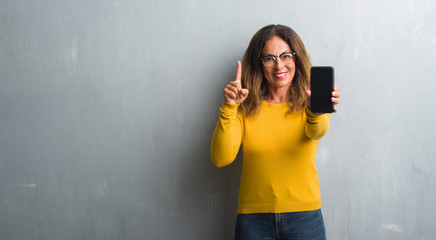 Middle age hispanic woman showing smarphone screen surprised with an idea or question pointing finger with happy face, number one