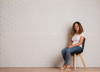 Middle age hispanic woman sitting on chair over white brick walll with a happy face standing and smiling with a confident smile showing teeth
