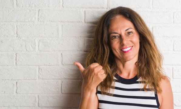 Middle Age Hispanic Woman Over White Brick Wall Pointing And Showing With Thumb Up To The Side With Happy Face Smiling