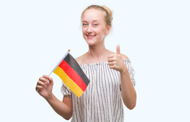 Blonde teenager woman holding flag of Germany happy with big smile doing ok sign, thumb up with fingers, excellent sign