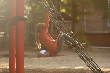 young blonde girl relaxing on a climbing net of a playground in a schoolyard in bright sunny light