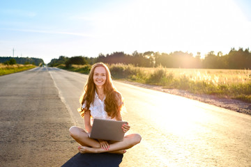 Beautiful young woman sitting with laptop and smiling on the road going beyond the horizon at sunset