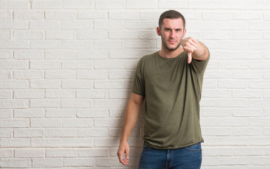 Young caucasian man standing over white brick wall looking unhappy and angry showing rejection and negative with thumbs down gesture. Bad expression.