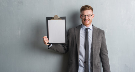 Young redhead business man over grey grunge wall holding clipboard with a happy face standing and smiling with a confident smile showing teeth