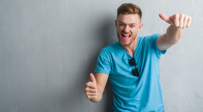 Young redhead man over grey grunge wall wearing casual outfit approving doing positive gesture with hand, thumbs up smiling and happy for success. Looking at the camera, winner gesture.