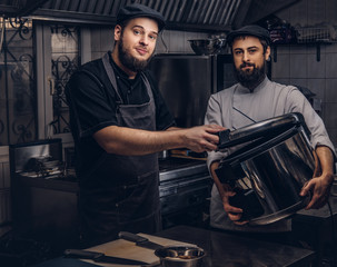 Two brutal cooks dressed in uniforms and hats standing with big multicooker in the kitchen.