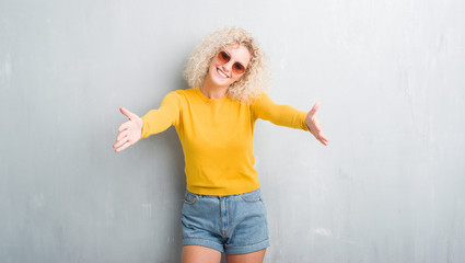 Young blonde woman with curly hair over grunge grey background looking at the camera smiling with open arms for hug. Cheerful expression embracing happiness.
