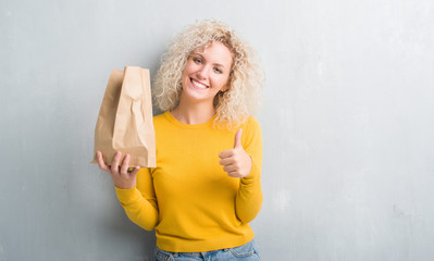 Young blonde woman over grunge grey background holding lunch paper bag happy with big smile doing ok sign, thumb up with fingers, excellent sign