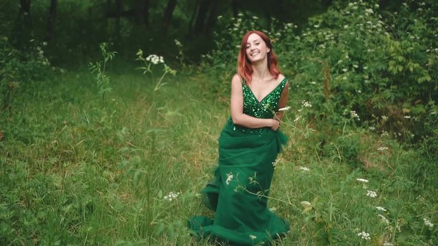 Red-haired girl with a hairstyle of quads, in a luxurious green dress with a long train. The princess stands in the midst of a forest glade and merrily waves her skirt with a hem. Slow motion