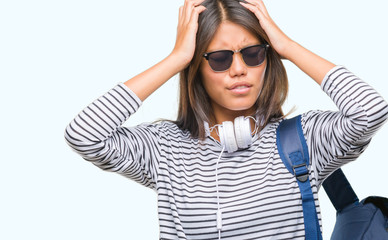 Young asian student woman wearing headphones and backpack over isolated background suffering from headache desperate and stressed because pain and migraine. Hands on head.