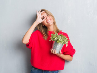 Beautiful young woman over grunge grey wall holding plant pot with happy face smiling doing ok sign with hand on eye looking through fingers