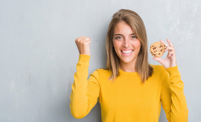 Beautiful young woman over grunge grey wall eating chocolate chip cooky screaming proud and celebrating victory and success very excited, cheering emotion