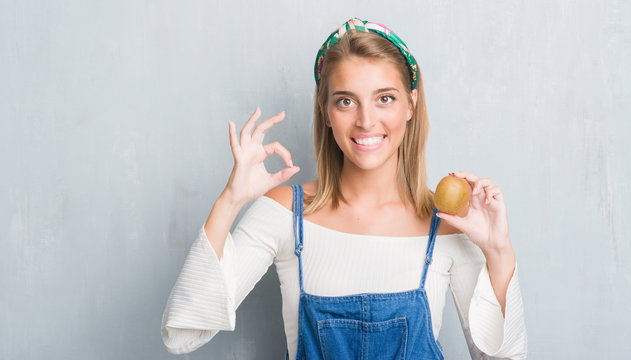 Beautiful young woman over grunge grey wall holding fresh kiwi doing ok sign with fingers, excellent symbol